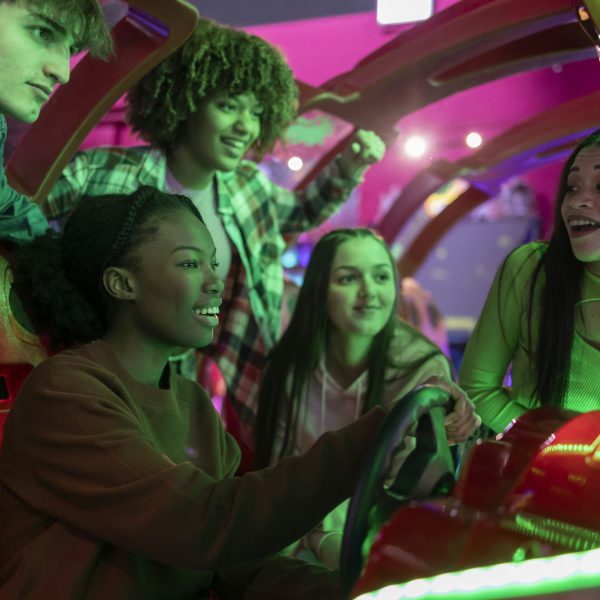 group of multiracial teenage friends in arcade playing car games with steering wheel